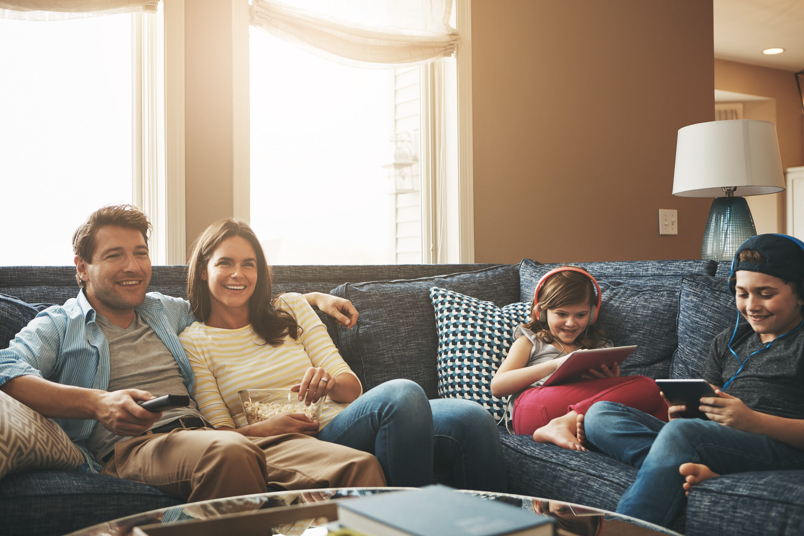 Technology, happy family together and in living room of their home with lens flare. Connectivity, networking or streaming and parents watching tv with children on tablet or smartphone with headphones.